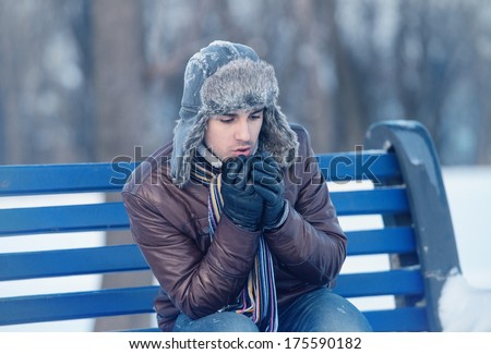 Winter walk - young man in an ear flap hat sitting on a bench and warming his hands