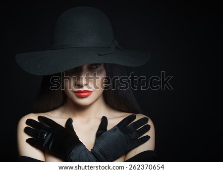 Woman in big black hat with arms across, studio portrait, dark background
