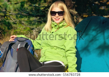 Young Woman with Smiling Face Hiker sitting with backpack and Tent Camping Outdoor on Grass with forest nature on background Travel and Healthy Lifestyle concept