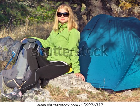 Young Woman with Smiling Face Hiker sitting with backpack and Tent Camping Outdoor on Grass with forest nature on background Travel and Healthy Lifestyle concept