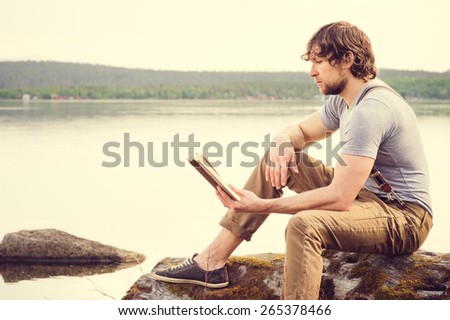 Young Man reading book outdoor with scandinavian lake on background Education and Lifestyle Travel concept