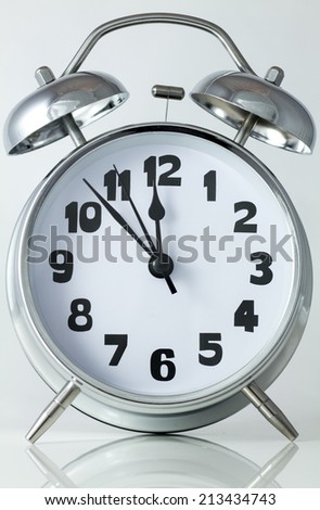 old style silver alarm clock with hammer and bells on top with hands at 7 minutes to midnight
