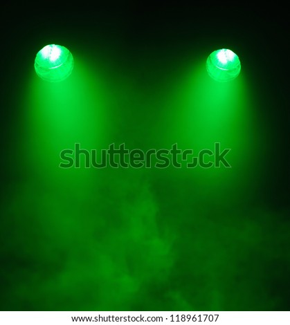 Two converging angled broad beamed green spotlights shining through a very smoky atmosphere in darkness with the electrical elements visible in the lights