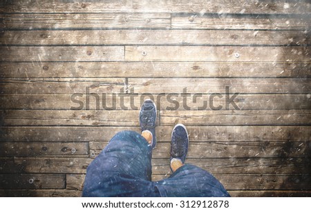 A man walking on rainy autumn season day on aged wooden floor, point of view perspective. Heavy rain and a man with blue shoes and shorts jeans walking alone on old wooden bridge. Conceptual photo.
