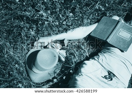 Young woman body parts with hat and old book lying in the grass. A woman thinking about book and enjoy. Blue cyan color filter effect used.