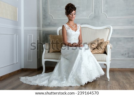Portrait of young luxurious brunette bride sitting on vintage sofa in beautiful wedding dress