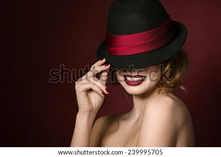 Portrait of young beautiful actress holding black hat with red ribbon over dark red background