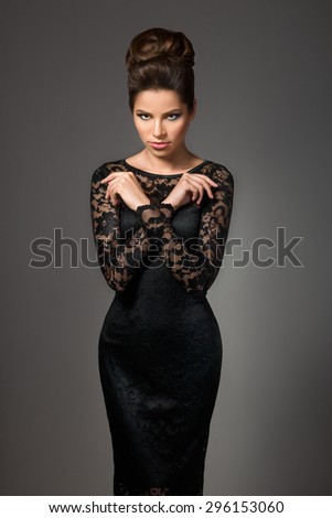 Beautiful young model in black dress with evening makeup and hairdo posing over gray background