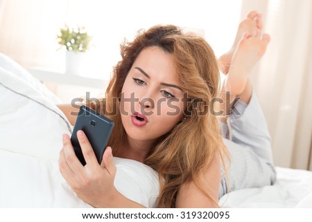 Young beautiful woman laying in her bed and looking at mobile phone. Morning time. Shocking news or getting late concept.