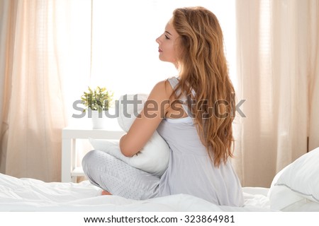 Young beautiful pensive woman sitting on her bed and holding her pillow. Calm sunny morning.