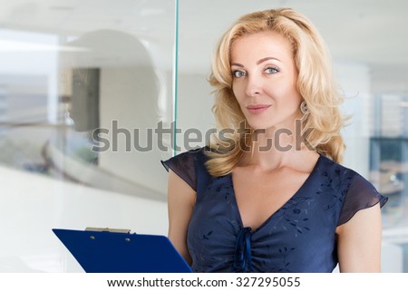 Portrait of beautiful adult business woman standing at her office and holding blue clipping pad with documents. Copyspace
