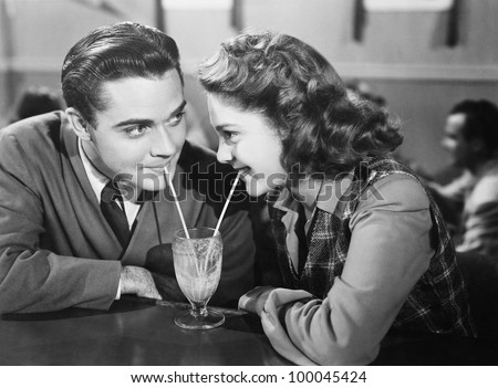 Couple In A Restaurant Looking At Each Other And Sharing A Milk Shake With Two Straws
