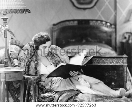 Young woman reclining in an armchair and reading a book in her bed room