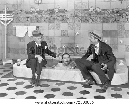 Two men sitting at the edge of a bathtub in suits and scrubbing a friends back