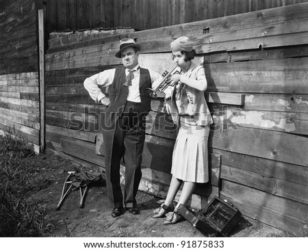 Woman practicing her trumpet while a man is standing next to her looking annoyed