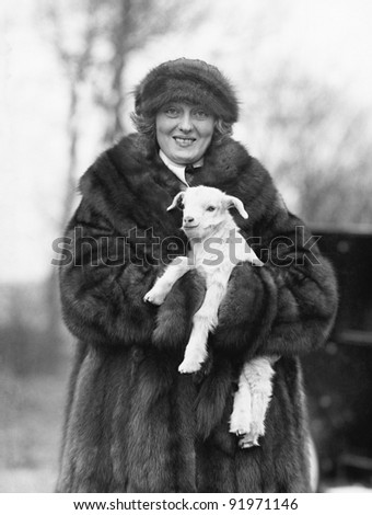 Woman in a fur coat and hat holding a small baby lamb in her arms