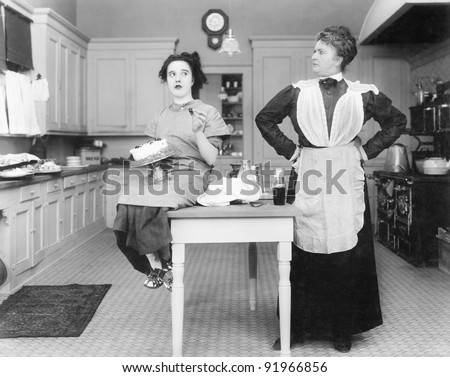 Housekeeper in the kitchen glaring at a young woman eating a cake