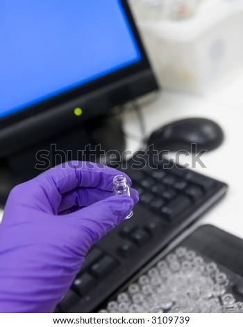 Science and computers. Gloved hand of scientist holding up a vial with a computer terminal in the background.  Shallow depth of field focus on the vial