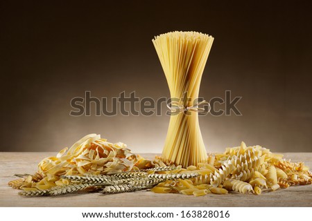 variety of uncooked pasta on wooden table with ears of wheat