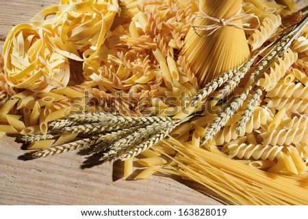 variety of uncooked pasta on wooden table with ears of wheat