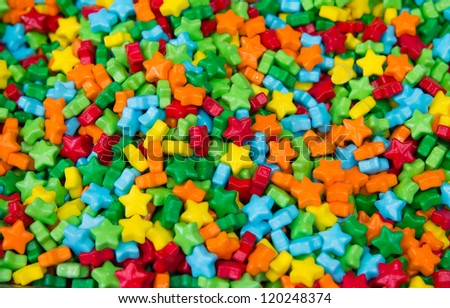 An assortment of shiny, brightly colored hard candy-coated stars which have a plastic look & feel to them fills the entire frame with focus at the center of the image & falling away towards the edges