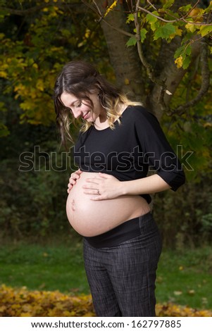 A young pregnant woman stands outside under an autumn tree. She is wearing a black shirt and her hands are on top of her bare pregnant and full belly.