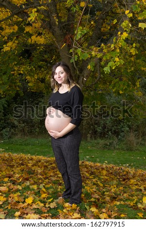 A young pregnant woman stands outside under an autumn tree. She is wearing a black shirt and her hands are on top of her bare pregnant and full belly.