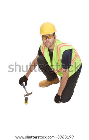 A construction worker is kneeling to pick up his claw foot hammer.  His tape measure is resting next to him.  He is wearing appropriate safety gear.