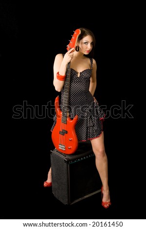 A beautiful brunette straddles a bass amplifier while holding a red electric bass. She is wearing a black & white polka dot dress, with red shoes & red painted nails.