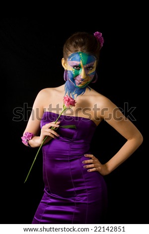 A woman with extreme full-face body painting poses on a black background holding a pink long stemmed rose. Her hand is on her hip and she is smiling. Mardi Gras color theme.