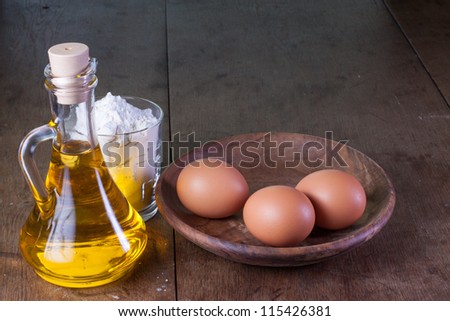 Oil in bottle, eggs in wooden plate, flour in glass on old oak table