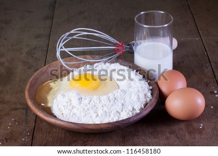 Broken egg, glass of milk and flour in wooden plate on old oak table