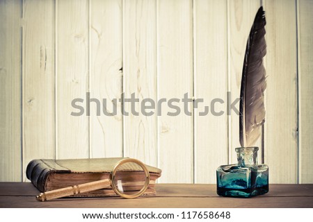 Quill pen, inkwell, book, magnifying glass on the table in front of wooden background