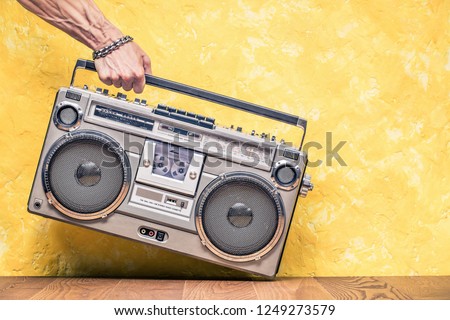 Retro outdated portable stereo boombox radio receiver with cassette recorder from circa 1980s in a strong man\'s hand front concrete textured yellow wall background. Vintage old style filtered photo