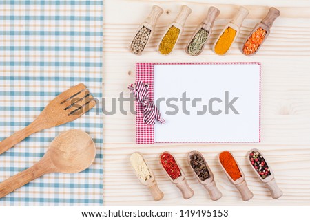 Recipe cooking book, spices in scoops, spoon, fork, tablecloth on wood table background