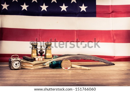 Vintage binoculars, quill pen, clock, compass, magnifying glass, old book, pocket knife, on table front USA flag background