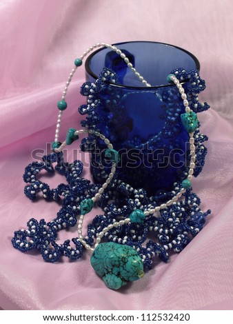 On a pink background there is a dark blue glass cup in  which there are necklaces from beads, pearls and a  turquoise stone