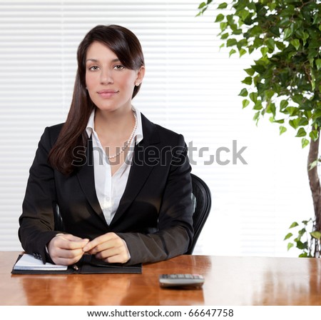 A woman in office attire/ business suit confidently sits behind her desk