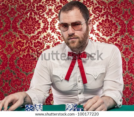 A man wearing glasses, a white shirt, and a red Texas tie sits at a blackjack table. There are stacks of blue and white chips in front of him./Confident Man Gambling Wearing Glasses, Texas Tie