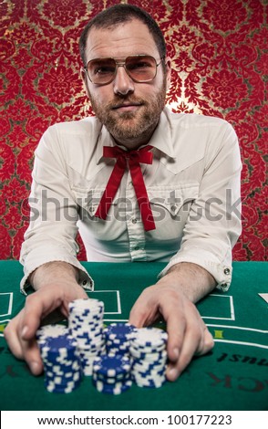 A man wearing glasses, a white shirt, and a red Texas tie sits at a blackjack table. He is making a big bet with all of his chips./Man Playing Blackjack Bets All His Money