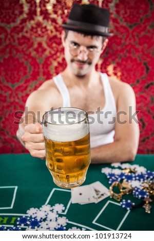 Mustache wife beater glasses man in hat toasting with glass of beer at vintage casino poker table