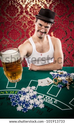 White wife beater guy in hat, glasses, mustache toasting friendly with large glass of light golden beer at Casino betting table