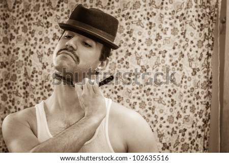 Young man, vintage sepia color, shaving neck in bathroom mirror with old straight razor, reflection, wearing wife beater, hat, shower curtain behind