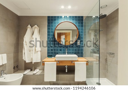 Little luxury bathroom with white bathrobes hanging on the wall. Round mirror, shower behind a glass wall. Gray wall blue tiles