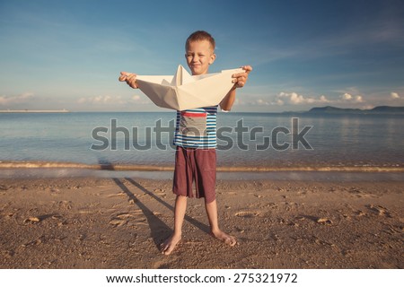 Happy kid playing with toy paper ship against blue summer sky and sea background.