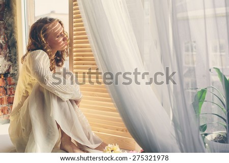 Beautiful young woman in a white dress sitting on the window in sunshine