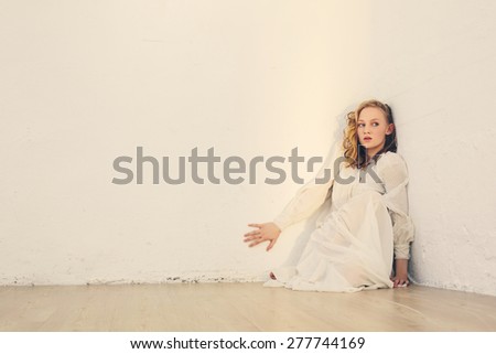 Scared young beautiful woman with curly hair sitting near white wall in the room