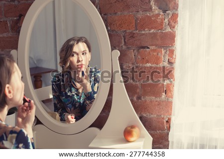 Beautiful woman doing make up and coloring her lips looking to the mirror in bedroom interior near a large window