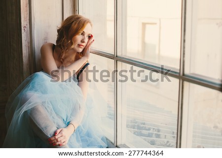 Toned portrait of a young red hair woman in blue fluffy skirt near the big window