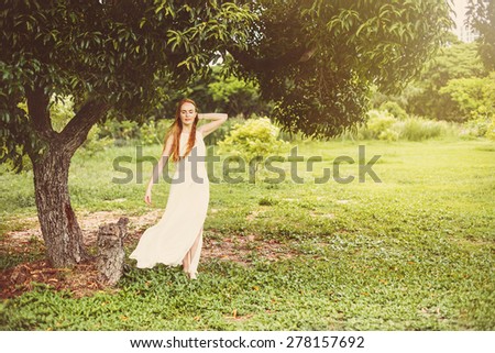 Beautiful young woman in long white dress with long red hair standing in wind under big tree on  green meadow